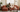 A couple relaxing on a rust-orange M1 modular sofa, one with a laptop and one reading, beside a record shelf